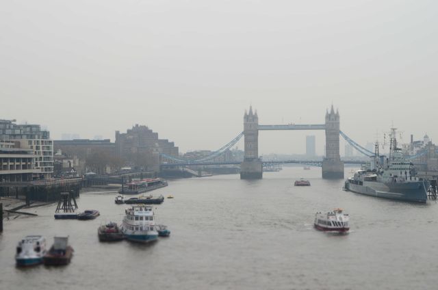 Depicting a foggy day over the river Thames with a view of Tower Bridge and several boats. The gray sky and mist create a moody atmosphere, highlighting the iconic and historic architecture of Tower Bridge. Useful for articles about London tourism, historic sites, and navigational waterways, also ideal as a background for travel-themed content.