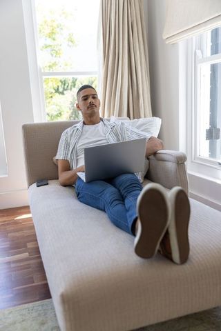 Relaxing mid-adult man working on laptop on chaise lounge in sunlit modern living room