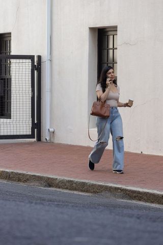 Woman walking on urban sidewalk talking on phone holding coffee and brown leather satchel