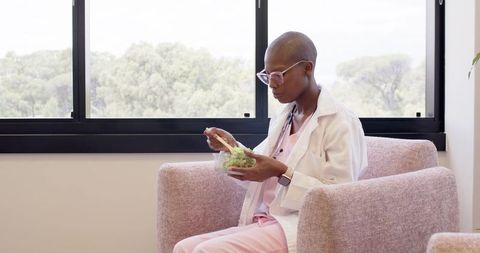 Clinician eating healthy salad in breakroom armchair wearing lab coat and smartwatch