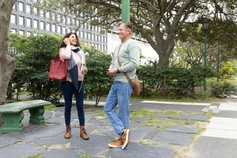Middle-aged couple smiling and chatting in city park holding white flower, smartphone, red tote