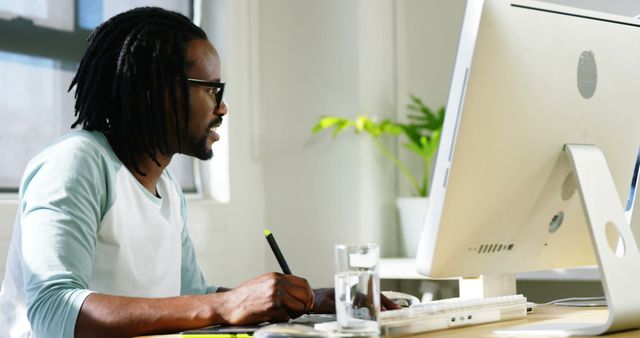 This image shows an African American man working at a desk in a bright, modern office. He wears casual attire and has dreadlocked hair. There is a computer, glass of water, and some papers on the desk, reflecting a productive working environment. Ideal for use in articles or advertisements about remote work, office productivity, modern workspaces, or diversity in the workplace.