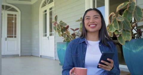 Smiling asian woman on porch enjoying coffee while using smartphone near potted plants