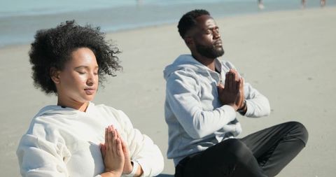 Diverse couple meditating on sunlit beach practicing mindfulness and breathing exercises