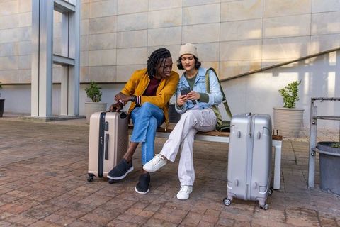 Diverse travelers sitting on bench checking smartphone with suitcases in urban plaza