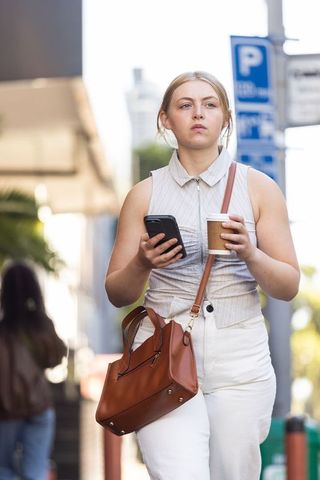Young woman walking downtown holding coffee and smartphone with leather crossbody bag