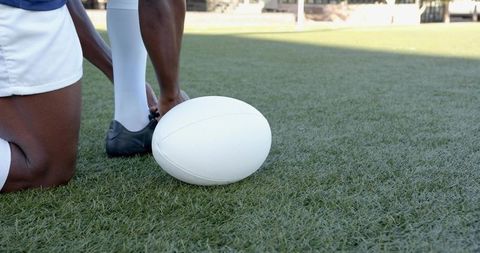 Kneeling athlete tying cleat beside white rugby ball on turf during pregame routine
