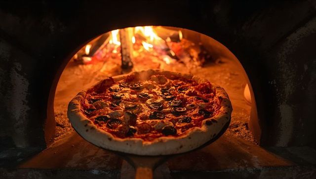 Baking wood-fired pepperoni pizza on peel at oven mouth with glowing flames