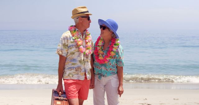 Senior couple dressed in summer attire, wearing leis, standing hand in hand by the ocean. Ideal for use in travel brochures, vacation advertisements, senior lifestyle promotions, or tropical holiday marketing.