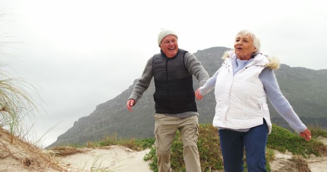 Senior couple holding hands walking on beach near mountains in winter clothing, enjoying a hiking adventure. Perfect for articles about active lifestyle and healthy aging, elderly fitness, retirement activities, enjoying nature, and love and companionship in later life.