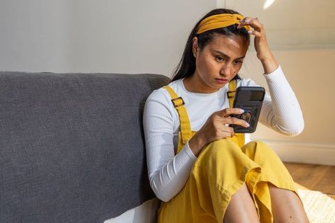 Asian woman sitting on living room floor in yellow overalls checking smartphone intently