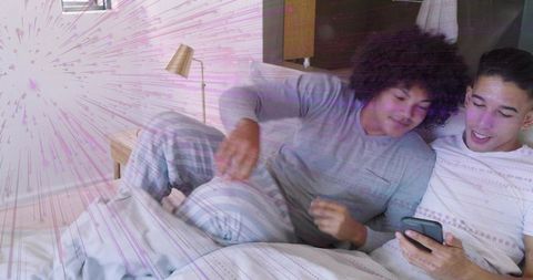 Two men relaxing in bed sharing smartphone, laughing in pajamas with minimalist lamp