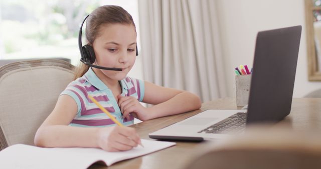 Young girl is using a laptop and wearing a headset while studying at home. She is taking notes and focusing on her schoolwork. Perfect for illustrating concepts of remote learning, online education, and home studies for children.