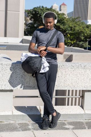 African american man leaning on rooftop checking smartwatch with duffel and headphones
