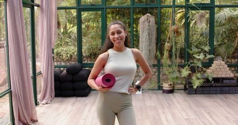 Smiling Woman Holding Yoga Mat in Bright Glass House Yoga Studio