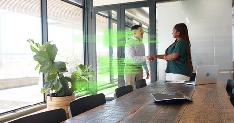 Business partners shaking hands across rustic wooden conference table in sunlit modern office