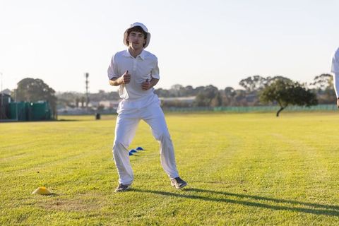 Young cricketer practicing fielding drill in white uniform and bucket hat on sunny grass