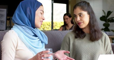 Indian women chatting on couch, hijab woman holding glass and friend gesturing by laptop