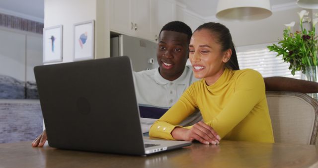 Image of happy diverse couple watching laptop together and smiling sitting in kitchen. Communication, happiness, love, domestic life and inclusivity concept.