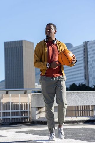 African american man standing on rooftop cradling basketball with mustard jacket and headphones