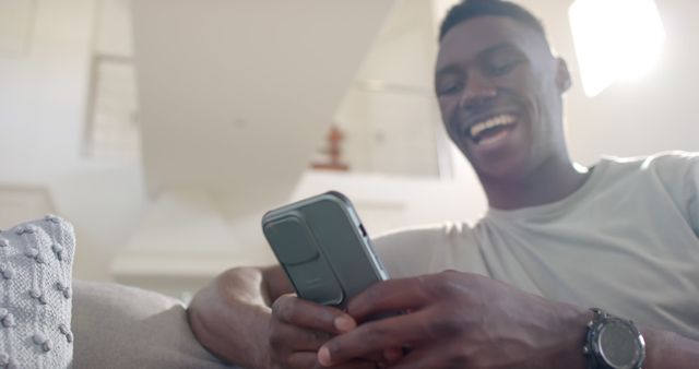 Cheerful man laughing while holding a smartphone in a modern living room. Ideal for themes related to communication, happiness, technology at home, or modern lifestyle.