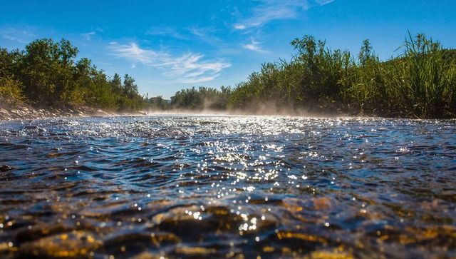 Sunlit shallow river rippling over pebble bed with morning mist glittering water surface