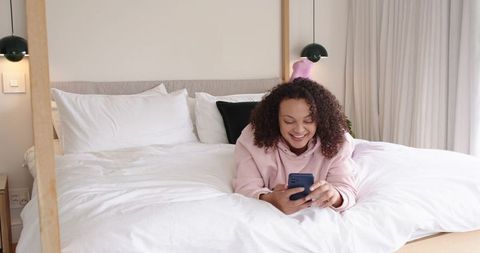 African american woman relaxing on white bed using smartphone in cozy minimalist bedroom