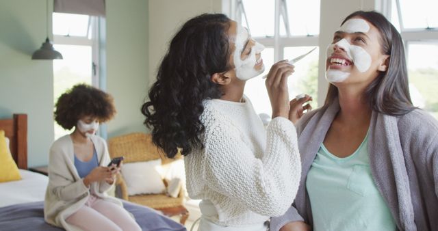 Young women are enjoying a spa day together indoors, applying face masks and sharing laughs. One woman is applying a face mask to her friend's face, while another is sitting on the bed using her phone. Perfect for use in content related to skincare, wellness, beauty routines, friendship, and self-care activities.