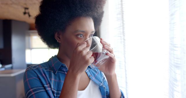 Woman with Afro hair stands by a sunlit window, savoring coffee in a modern home. She wears a casual blue plaid shirt and gazes thoughtfully outside. Perfect for use in blogs, websites, or advertisements focusing on lifestyle, relaxation, morning routines, self-care, and home settings.