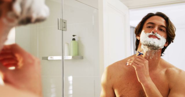 Shirtless man applying shaving cream and shaving facial hair with a razor in front of bathroom mirror. Perfect for advertisements, articles, or blogs related to male grooming, personal care products, morning routines, hygiene tips, or self-care education.