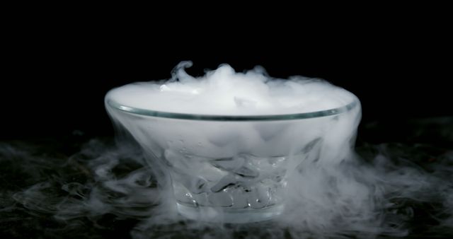 A glass bowl is filled with a substance that is emitting smoke or vapor, creating a mysterious and dramatic effect. The smoke swirls around the bowl, suggesting a chemical or culinary experiment.