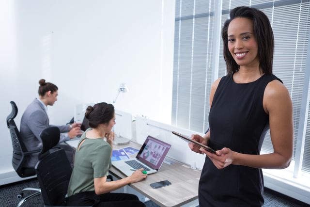 Confident female executive holding tablet and smiling in a modern office with colleagues working in the background. Perfect for illustrating business, technology in the workplace, professional environments, and teamwork.