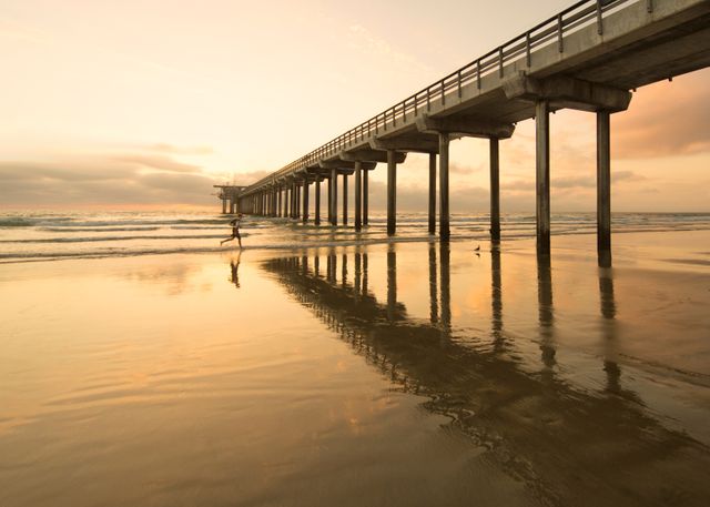 Pier extending into ocean during beautiful sunset, casting a warm glow on sand and water. Silhouette of a person walking under pier. Ideal for travel, tourism promotions, beach themes, and relaxing atmosphere projects.