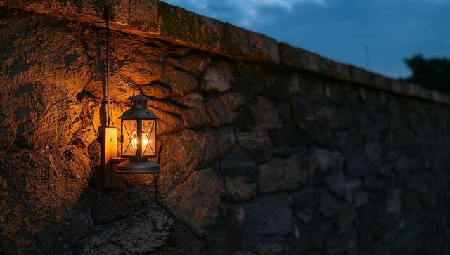 Glowing lantern casting warm amber light on rough stone wall at dusk with vintage bulb