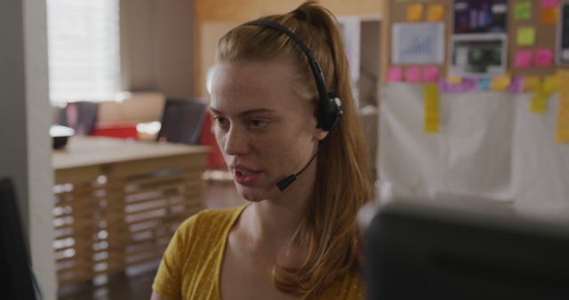 Young woman with a headset working at a computer in a modern office environment. Background includes whiteboards and charts with ideas and strategies typical in collaborative workspaces, suggesting a focus on productivity and teamwork. Ideal for use in business, technology, and communication themed projects, as well as showcasing modern workspaces and professional environments.