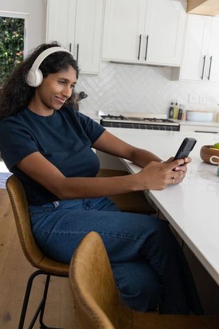 Relaxing african american woman listening with headphones and using smartphone at kitchen island