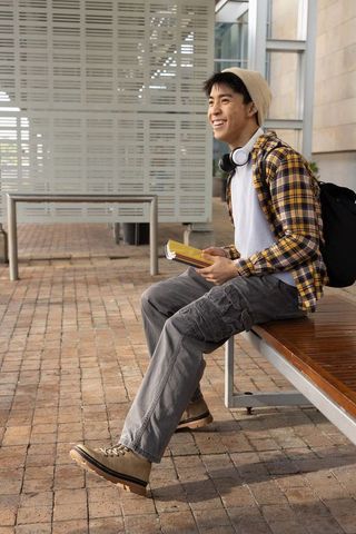 Smiling asian college student sitting on bench studying books, headphones and backpack