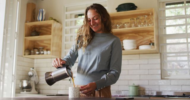 Smiling woman pouring coffee from French press into cup in a modern kitchen with wooden shelves and white countertops. Might be used for illustrating domestic lifestyles, morning routines, breakfast habits, and home interiors. Ideal for content related to hospitality, recipes, home improvement, or kitchen appliances.