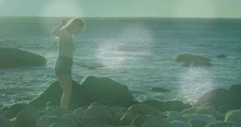 Young woman adjusting hair on rocky shore at golden hour, barefoot in denim shorts, lens flare