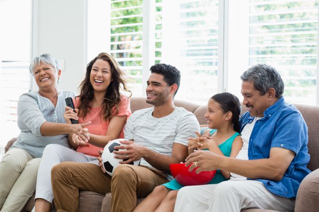 Multi-generation family enjoying time together watching a soccer match on television in a bright living room. Ideal for use in advertisements promoting family bonding, leisure activities, sports events, and home entertainment products.