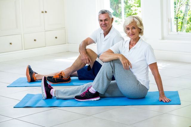 Senior couple sitting on yoga mats in a bright room, engaging in stretching exercises. Both are dressed in comfortable athletic wear, smiling and looking at the camera. Ideal for use in articles or advertisements promoting senior fitness, healthy aging, home workouts, and wellness programs for elderly individuals.