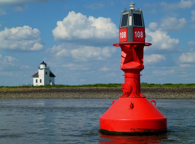Red buoy gently floating on calm water under partly cloudy blue sky with a lighthouse in the background. Ideal for illustrating nautical themes, ocean safety, navigation aids, or coastal scenery.