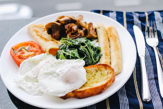 Delicious and balanced breakfast plate featuring poached eggs, grilled sausages, freshness of grilled tomato, sautéed spinach, and mushrooms, served with toast. Ideal for use in nutrition blogs, healthy eating campaigns, restaurant menus, and breakfast-themed advertisements. Showcases importance of a balanced meal for a healthy start to the day.
