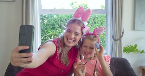 Mother and daughter taking easter selfie with bunny ears and painted egg during spring home celebrat