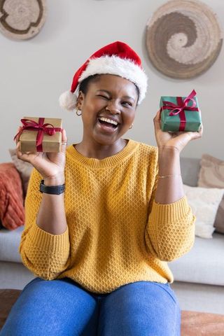 African american woman wearing santa hat holding two gifts, winking and laughing in cozy living room