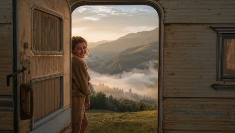 Smiling woman standing in camper doorway looking back at misty mountain valley at sunrise