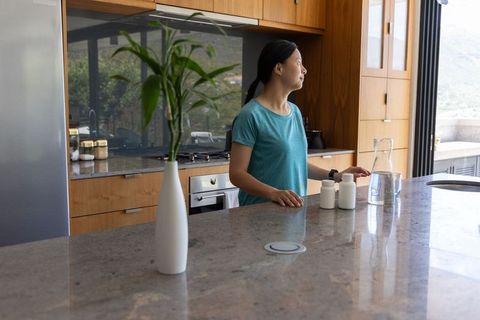 Asian woman standing in modern kitchen touching counter near pill bottles and glass carafe