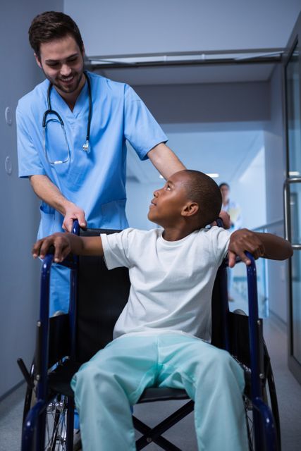 Male nurse in blue scrubs interacting with a child patient in a wheelchair in a hospital corridor. The nurse is smiling and engaging with the child, creating a comforting and supportive atmosphere. This image can be used for healthcare, medical, and pediatric care themes, illustrating patient care, nursing, and hospital environments.