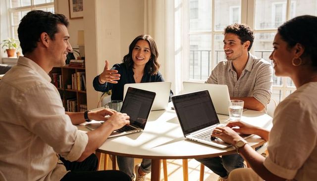 Young professionals collaborating at laptop-filled round table in bright coworking studio