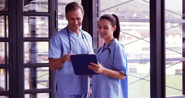 This image depicts two healthcare professionals in blue scrubs collaborating in a modern hospital setting. They are engaged in a discussion while referring to a clipboard, suggesting planning or reviewing of patient care. Ideal for topics related to medical teamwork, healthcare communication, hospital environment, medical consultations, and staff coordination.